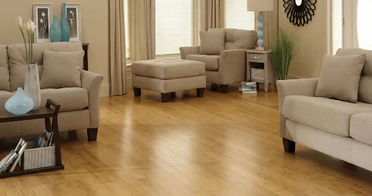A living room with professionally installed bamboo flooring, beige sofas, a matching ottoman, a side table with a blue lamp, vases, framed pictures, and large windows with blinds and curtains in Salt Lake City, UT